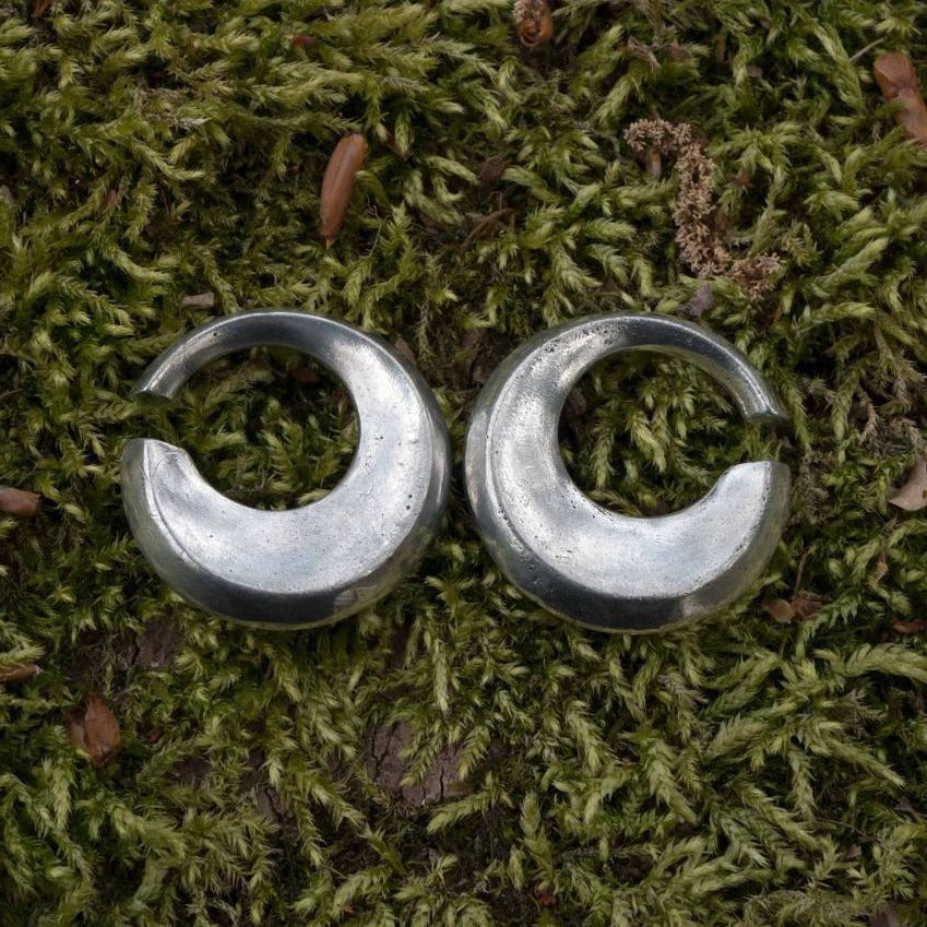 Two circular HOLO Hoop Dangle Ear Weights in Silver by Kaya Movement, with an opening on one side resembling broken rings, are placed side by side on a bed of green moss. The slightly tarnished surface of these 6 gauge earrings suggests an old or rustic style of body jewelry.
